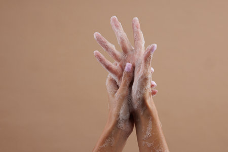 A close-up of two hands lathered with soap, demonstrating hand washing. The hands are positioned in a way that emphasizes the soap bubbles against a neutral brown background.の写真素材