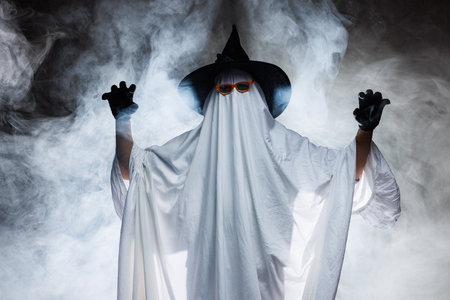 Man in scary ghost costume for Halloween and witch hat on dark mystical background in smoke, Concept of Halloweenの写真素材