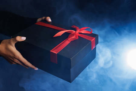 A person holding a black gift box with a red ribbon against a smoky blue background, creating a...