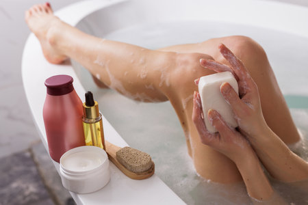 woman holding a bar of soap while sitting in a bathtub with foam, body and hair care products lying nearby,の写真素材