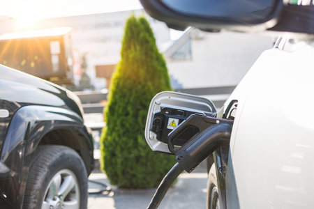 An electric vehicle charging at a station, with a close-up of the charging port. In the background, another car is parked, and a green shrub is visible, illuminated by sunlight.の写真素材