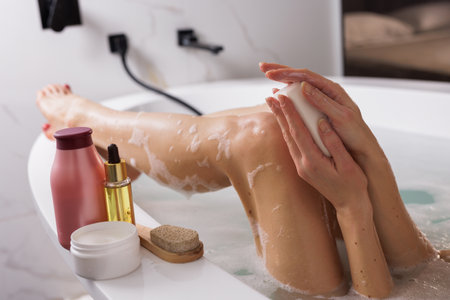 Relaxing bath time: Woman enjoying a self-care ritual in a bathtub, lathering soap on her hands. Spa essentials like body lotion, oil, and a pumice stone create a soothing wellness atmosphereの写真素材