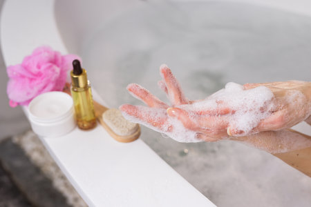 Relaxing spa moment. Woman washing hands with foamy soap near a bathtub. Skincare essentials like...