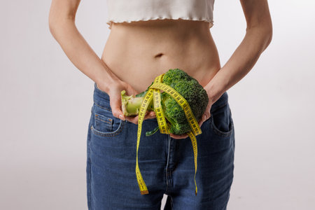 Slim woman holding broccoli wrapped in a measuring tape, symbolizing healthy eating, weight loss, diet, and nutrition on a white background.の写真素材