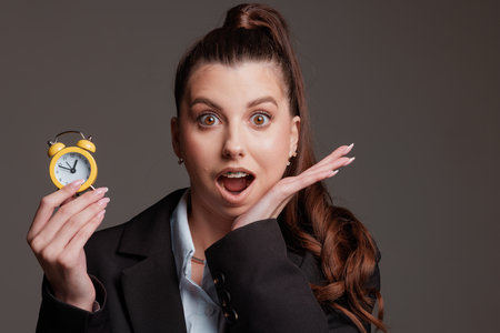 Surprised businesswoman holding a small yellow alarm clock, expressing shock at the time. Concept of time management, deadlines, and urgencyの写真素材