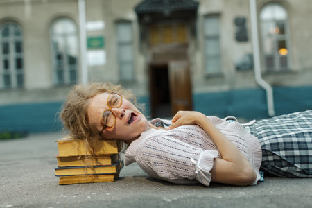 Nerdy young woman in oversized glasses struggles to carry heavy books, yawning or gasping in exhaustion while standing outdoors in sunlight.の写真素材