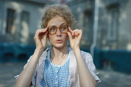 Young woman with freckles adjusting oversized vintage glasses, looking curiously at the camera, wearing a knitted sweater and checkered dress outdoors.の写真素材