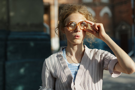 Young woman with messy curly hair and oversized vintage glasses looks surprised and confused,...