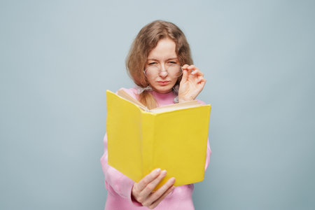 Serious woman in glasses reading yellow book, adjusting glasses, concentrated, wearing pink sweater,...
