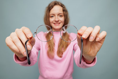A smiling woman in a pink sweater holds a pair of round glasses towards the camera, set against a light blue background.の写真素材