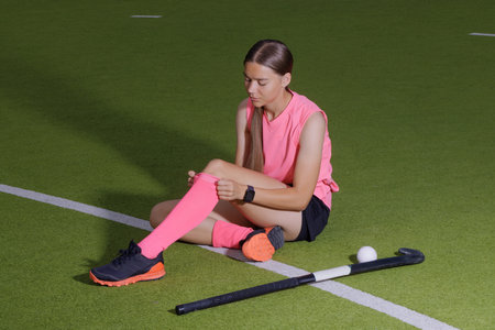 Young female field hockey player sitting on turf, adjusting socks before game, stick and ball nearby.の写真素材
