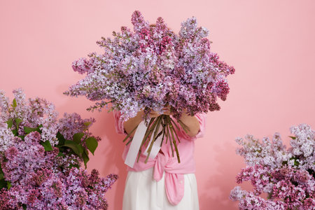 Woman hiding face behind large bouquet of blooming lilacs on pink background, spring floral concept.の写真素材