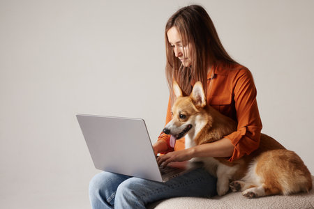 Young woman working remotely with her Corgi dog sitting on lap, using laptop at cozy home office.の写真素材