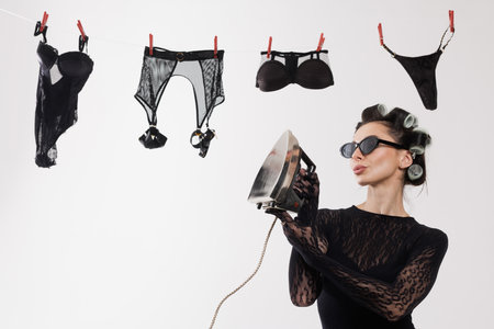 Glamorous woman with curlers holding iron near lingerie on clothesline in playful, retro-inspired domestic setting.の写真素材