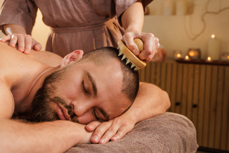 Male client experiences calming scalp massage using a brush tool in a peaceful spa. Promotes relaxation and head tension relief.の写真素材