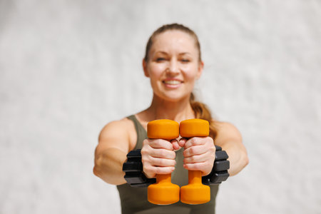 Fit woman smiling and holding orange dumbbells outdoors, showcasing motivation, strength, and...