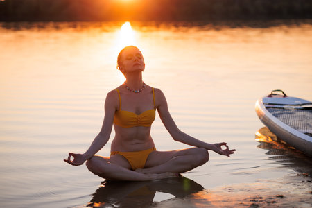 Peaceful woman practicing meditation at sunset by the water in a lotus pose, embracing calmness, nature, and mindful awareness near paddleboard.の写真素材
