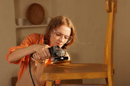 Woman using electric sander on wooden chair, home furniture restoration, repair and sanding process.