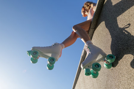 Low angle shot of woman wearing retro roller skates sitting on wall against blue sky summer outdoor lifestyle with copy spaceの写真素材