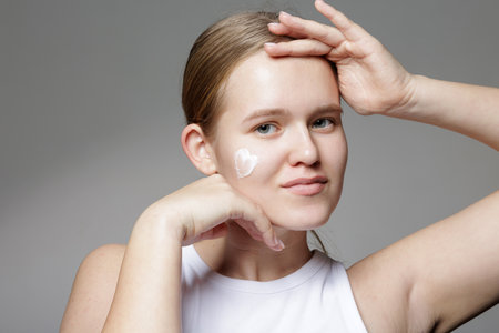 Smiling young woman with moisturizing cream spot on cheek poses on gray background, illustrating soft daily facial care and healthy skin.の写真素材