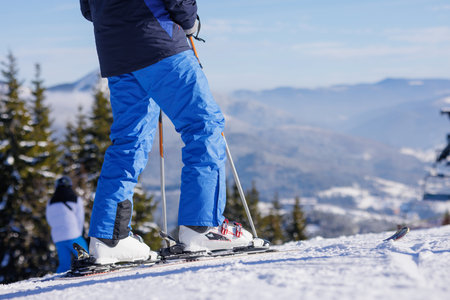 Low angle close up of skier legs and skis on snowy slope with alpine mountains and winter resort backgroundの写真素材