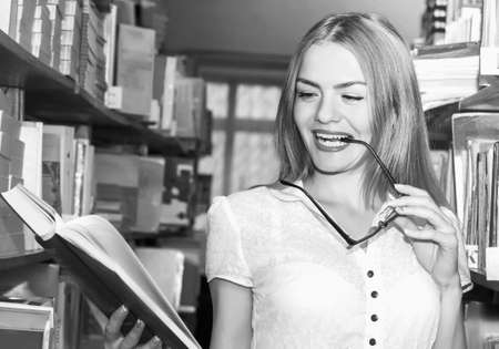 GIRL IN THE LIBRARY BETWEEN racks with books, a beautiful blonde with glasses gets knowledge and education, the teaching professionの写真素材