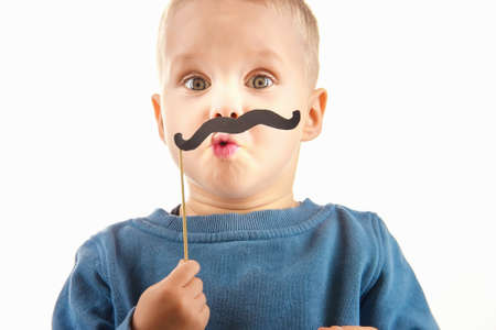 Happy little boy playing with a paper mustache, laughing while doing face, on white background, fun for childrenの写真素材