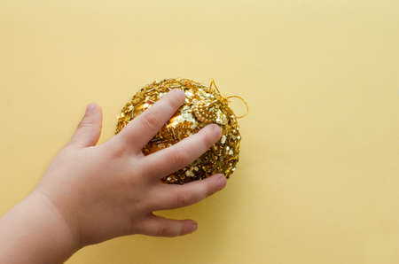 childrens hand puts golden christmas tree toy ball on yellow background, happy new year