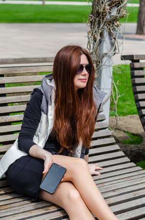 beautiful slender young white girl in sunglasses sits in the park with phoneの写真素材