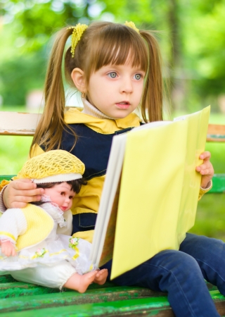 pretty young girl reads a book with her doll at the parkの写真素材
