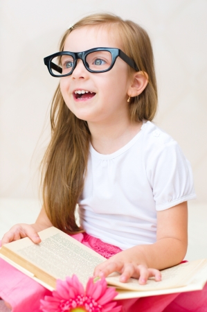 happy little girl is reading a book while wearing glasses indoorsの写真素材