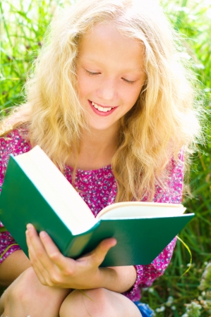 Young blond blue-eyed teenage girl sitting on the grass and reading a bookの写真素材