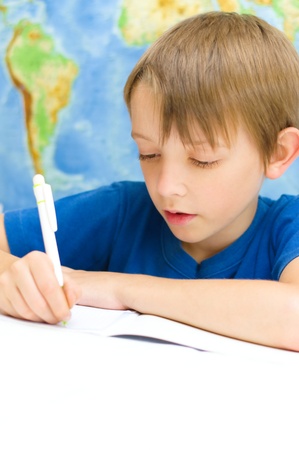 schoolboy writing in his workbook using a pen in front of world mapの写真素材