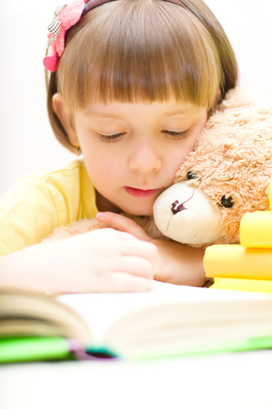Cute little girl with her toy bear are reading a book while sitting at tableの写真素材