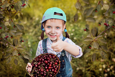 cute little girl with cherries showing her thumb up in the garden