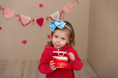 Little cute girl on the background wall of a house. The child in the scenery in the form of hearts. The kid drinks juice. Girl in a blue wrap and red dress drinks from a drink tubeの写真素材