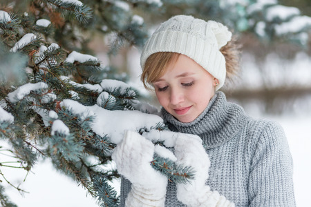Beautiful girl in a white hat and mittens in the winter snowy forest. Cute child in a knitted dress near a tree in the winterの写真素材