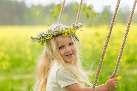 Beautiful little girl with a wreath on her head playing in a flowering field. A girl is playing in nature surrounded by yellow flowers. A young blonde is riding on a swing behind an open-air cageの写真素材
