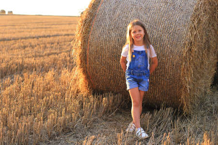 Beautiful little girl stands near a haystack in a summer field