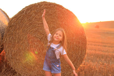 Beautiful little girl stands near a haystack in a summer fieldの写真素材