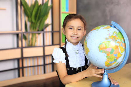 Portrait of a girl in a school uniform with a globe in her hands. Young schoolgirl in the class. The girl sits with a globe at her desk. Child at school.の写真素材