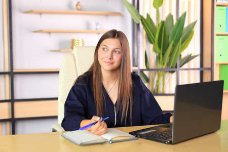 A young girl sits at a computer in the office. Schoolgirl in a laptop studies remotely. Office worker in an office with gadgets. Online training. Young teacher preparing for lessonsの写真素材