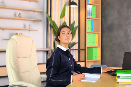A young girl sits at a computer in the office. Schoolgirl in a laptop studies remotely. Office worker in an office with gadgets. Online training. Young teacher preparing for lessonsの写真素材