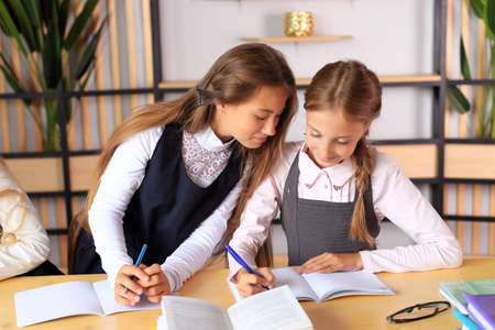 A young schoolgirl in a school uniform writes in a notebook. The child prepares lessons at the table. Girl writes in a notebook. Children in the classroom. School collectiveの写真素材