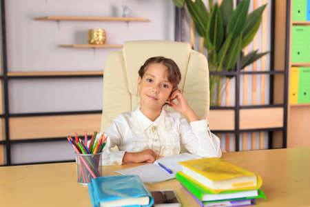 A young schoolgirl in a school uniform writes in a notebook. The child prepares lessons at the table. Girl writes in a notebookの写真素材