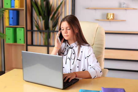 A young girl sits at a computer in the office. Schoolgirl in a laptop studies remotely. Office worker in an office with gadgets. Online training. Young teacher preparing for lessonsの写真素材