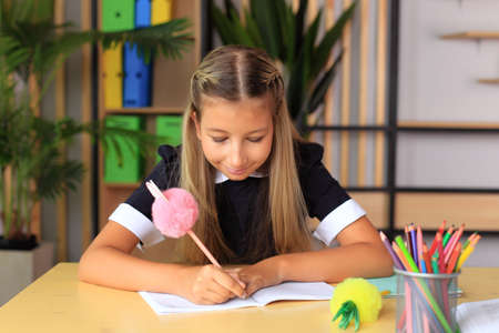 A young schoolgirl in a school uniform writes in a notebook. The child prepares lessons at the...