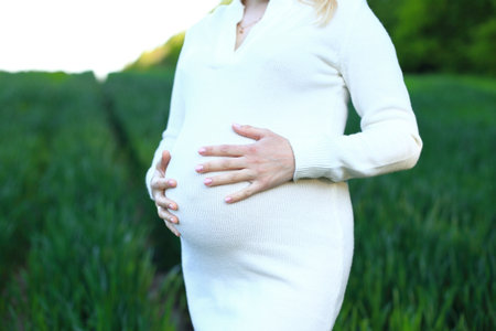 A pregnancy woman standing in a field, holding a bouquet of flowers in her hands. It is evident that she is embracing the moment and eagerly anticipating the arrival of her third child. The image captures a serene and joyous moment in which the woman is connected to the natural environment and celebrating the upcoming addition to her family. She is wearing a white dress and has a bright smile on her face, radiating happiness and contentment. The field is filled with lush green grass, and the sky above is a beautiful shade of blue. The serene and picturesque backdrop adds to the overall tranquility of the scene. The womans pose and the way she delicately holds the flowers suggest a sense of appreciation for the beauty of nature and the simple joys of life.の写真素材