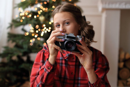 A girl in a striking red dress, holding a vintage camera in her hands. She sitting in front of a beautifully decorated Christmas tree, adorned with sparkling garlands and twinkling lightsの写真素材