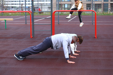 A father and his son engage in physical exercises, performing push-ups on a well-equipped outdoor...